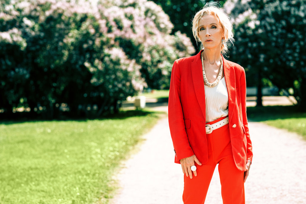 dans un jardin, une femme d'âge mûre porte un tailleur rouge avec des accessoires tel qu'un gros collier, une ceinture, une bague blancs.