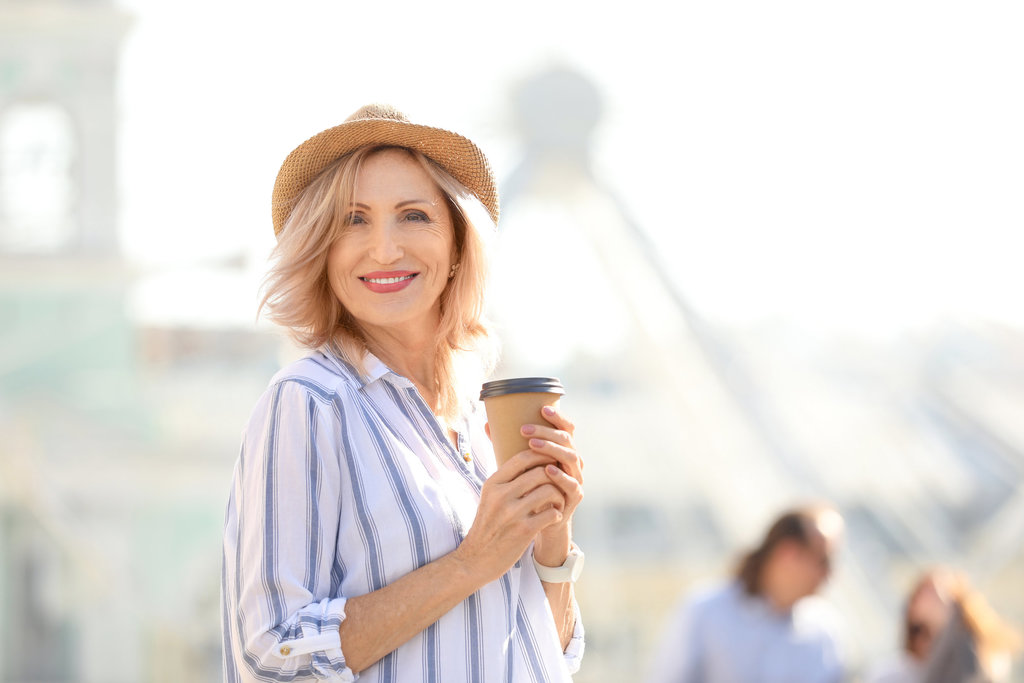 une femme de 50 ans prend la pose en tenant un café dans ses mains. elle porte une chemise blanche à rayures bleues verticales. elle porte sur la tête un chapeau. elle sourit à la photo.
