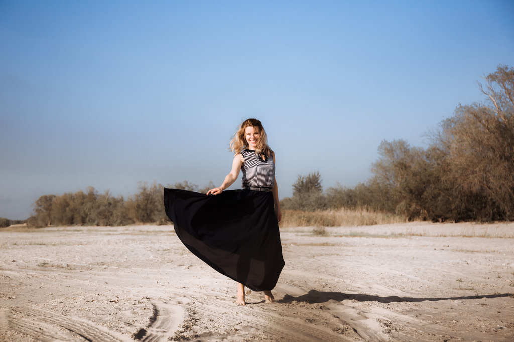 une jeune femme souriante soulève sa longue jupe noire, les pieds dans le sable.