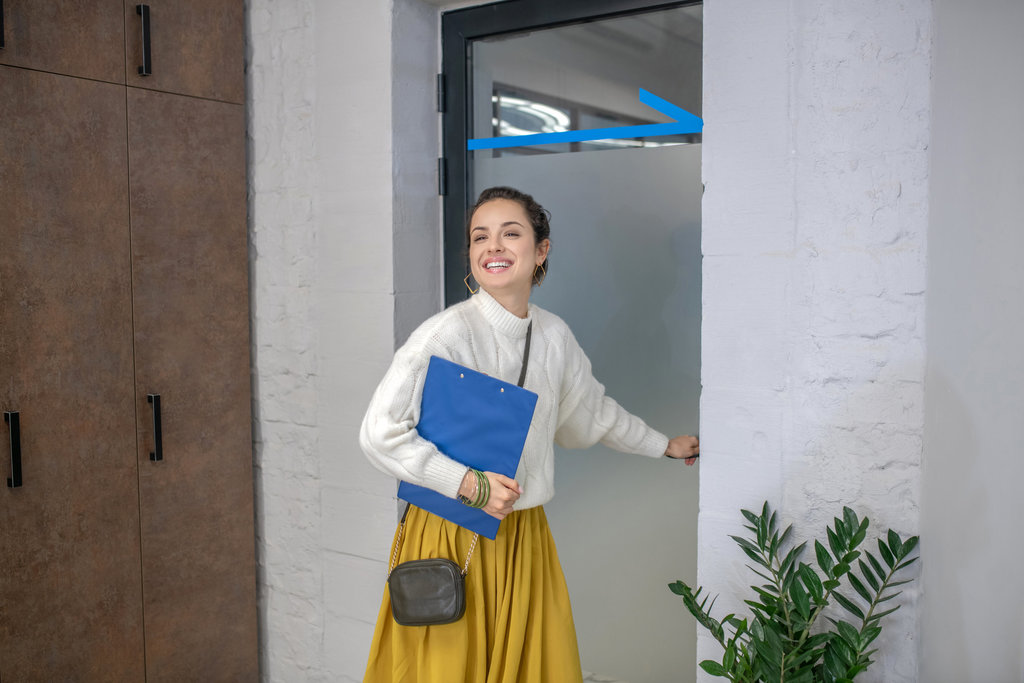 une jeune femme tient la porte d'un bureau d'une main et un chemisier dans l'autre maijn. Elle porte un pull blanc et une jupe longue jaune et sourit.