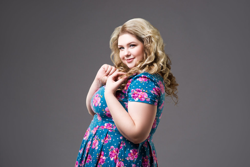 une jeune femme prend la pose de profil dans une jolie robe bleue à motifs fleuris rose. elle porte de jolie boucles blondes et sourit au photographe.