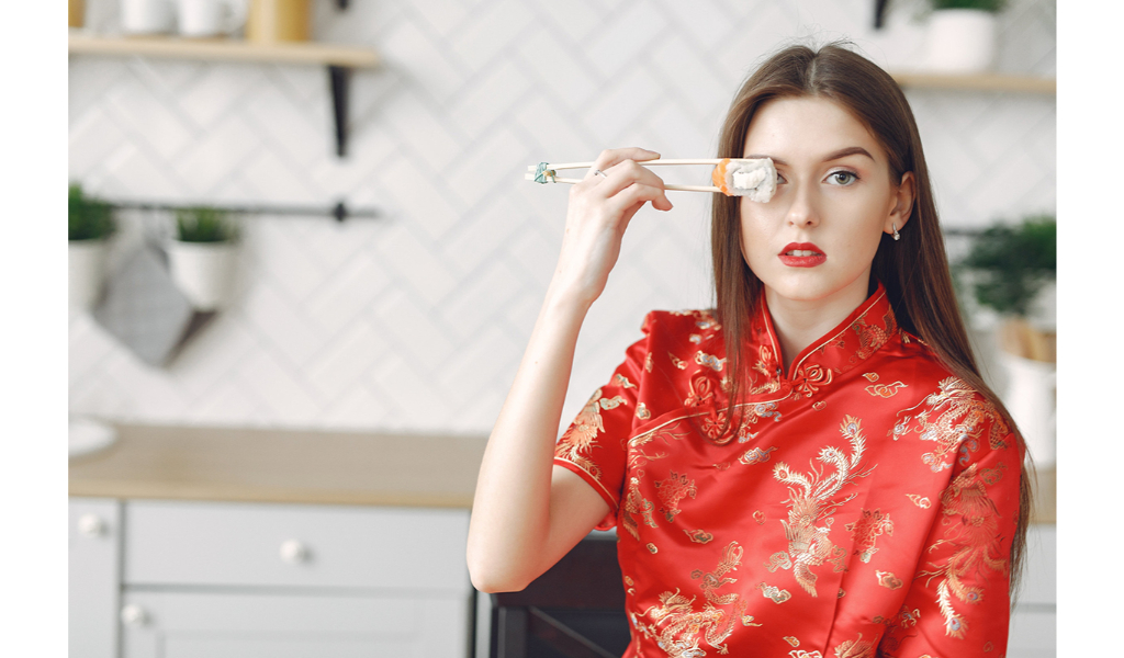 une jeune femme porte un kimono rouge avec des symboles dorés. elle est assise dans une cuisine et tient à la main des baguettes chinoises avec un sushi qu'elle met devant son oeil droit.
