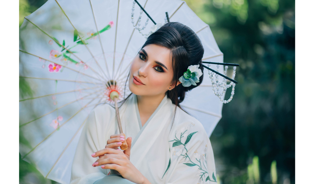 une jeune femme brune en kimono blanc regarde le photographe en tenant une ombrelle chinoise blanche avec des motifs de fleurs. elle porte des baguettes dans ses cheveux, avec des perles suspendues. elle a également une fleur blanche dans les cheveux.