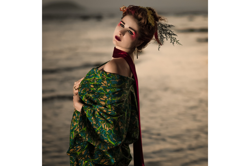 une asiatique en kimono prend la pose sur une plage de sable. elle porte un kimono à motifs vert, une écharpe rouge autour du cou et porte beaucoup de maquillage. Ses cheveux sont attachés.