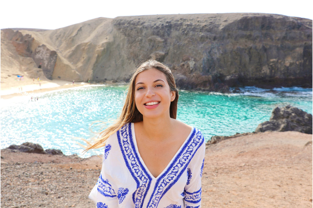 une jeune femme sourit dans une jolie tunique blanche et bleu avec un col en V. dans le fond voit une plage avec des falaises.