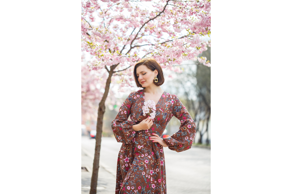une femme sous un cerisier rose en fleurs, porte un kimono robe de couleur marron avec des motifs de fleurs. elle tient une petite branche de cerisier dans sa main droite devant son buste.