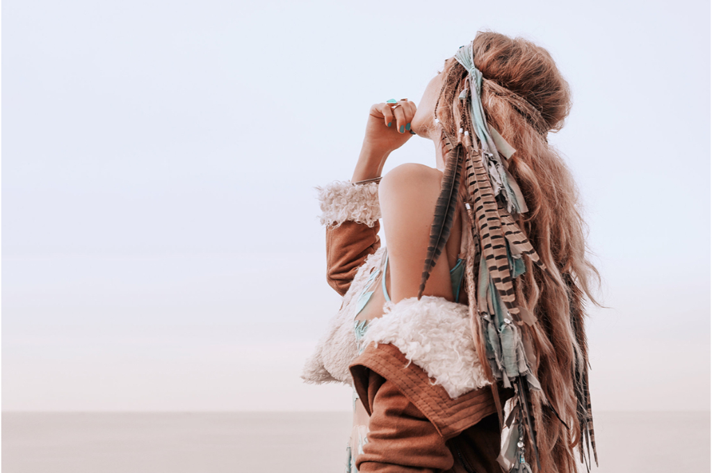 une femme de dos regarde en l'air dans le désert. ses cheveux sont attachés avec un headband avec divers éléments de tissu. elle porte un manteau en simlili cuir avec intérieur poils de mouton avec une épaule découverte.