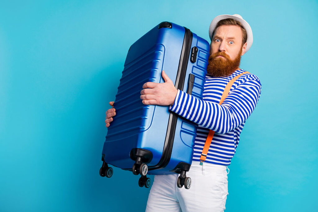 un homme porte entre ses bras une valise de voyage rigide bleue. il porte une marinière, ainsi qu'un jeans blanc avec des bretelles et un chapeau blanc. il est barbu aux yeux bleus et cheveux roux.