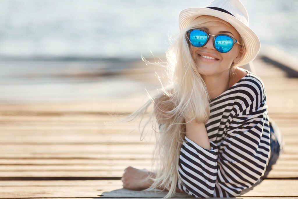 une jeune femme blonde est allongée sur un ponton en bois en direction du photographe. elle a de longs cheveux blonds sur lesquels est posé un chapeau. elle porte des lunettes de soleil et une marinière ample.