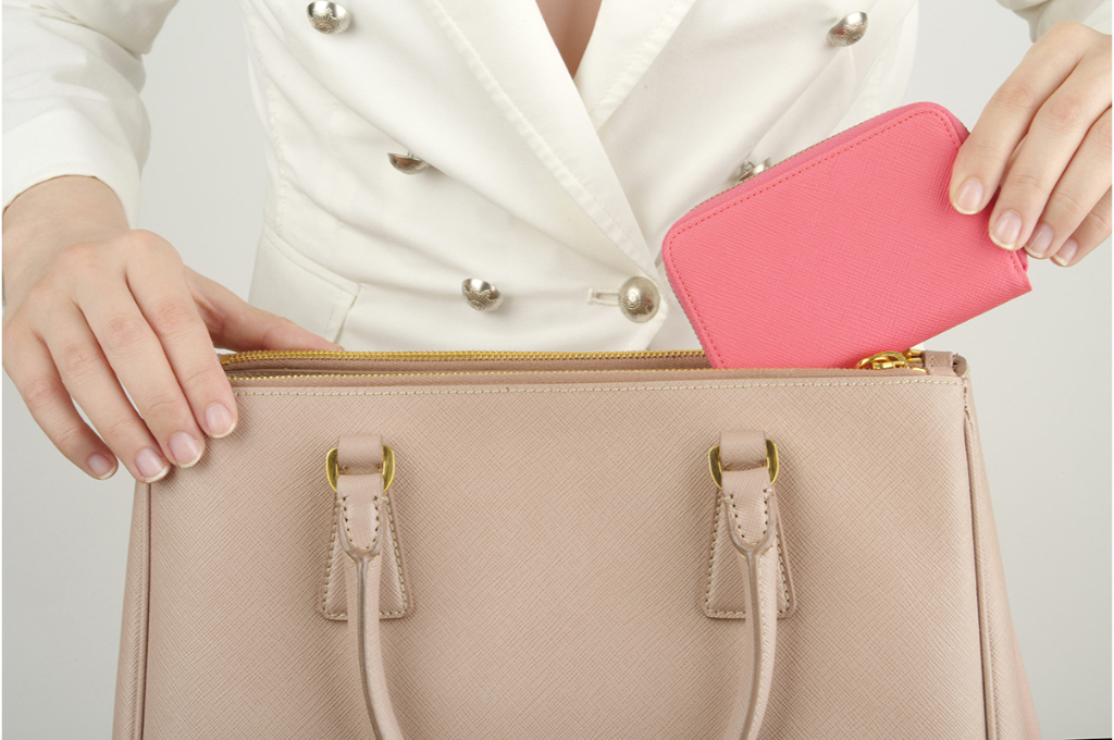 vue rapprochée sur un sac à main beige dans lequel une femme en veste blanche à boutons argentés, met un porte-monnaie rose à l'intérieur.