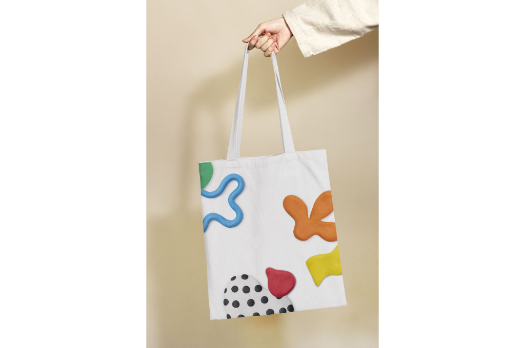 une main tient par la poignée un sac en tissu de créateur blanc avec des motifs de plusieurs couleurs.