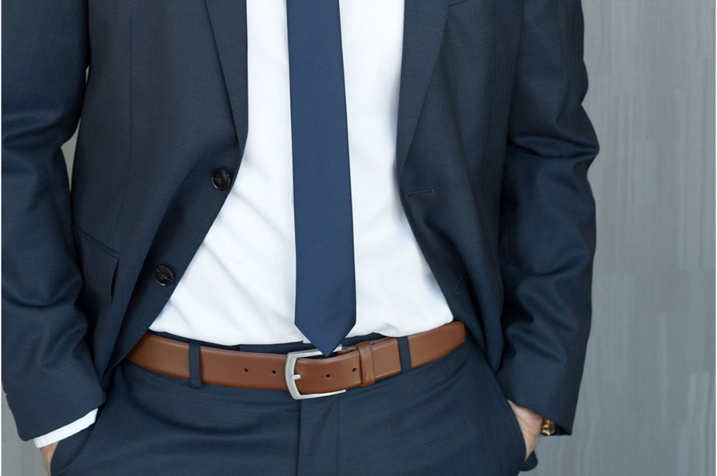 aperçu sur le buste d'un homme en costume qui a les mains dans les poches. son costume cravate est bleu marine avec une chemise blanche. il porte une ceinture marron au pantalon.