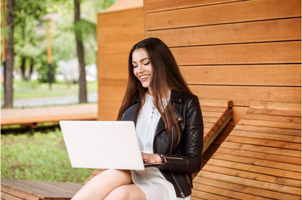 un jeune brune aux longs cheveux sourit, posée sur un transat en bois devant un chalet en bois. Elle a un ordinateur portable blanc posé sur ses genoux. Elle porte une veste en cuir sur une robe blanche.
