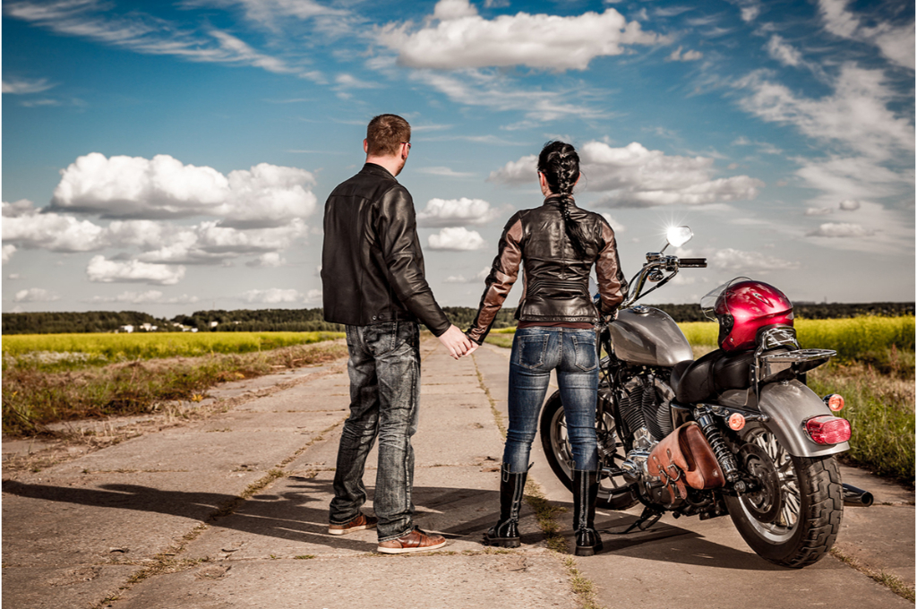 un couple de motards se tiennent la main. Ils sont dos à la photo et regardent ensemble la route aux côtés de leur moto. Ils portent tous les deux une veste en cuir et un jean.