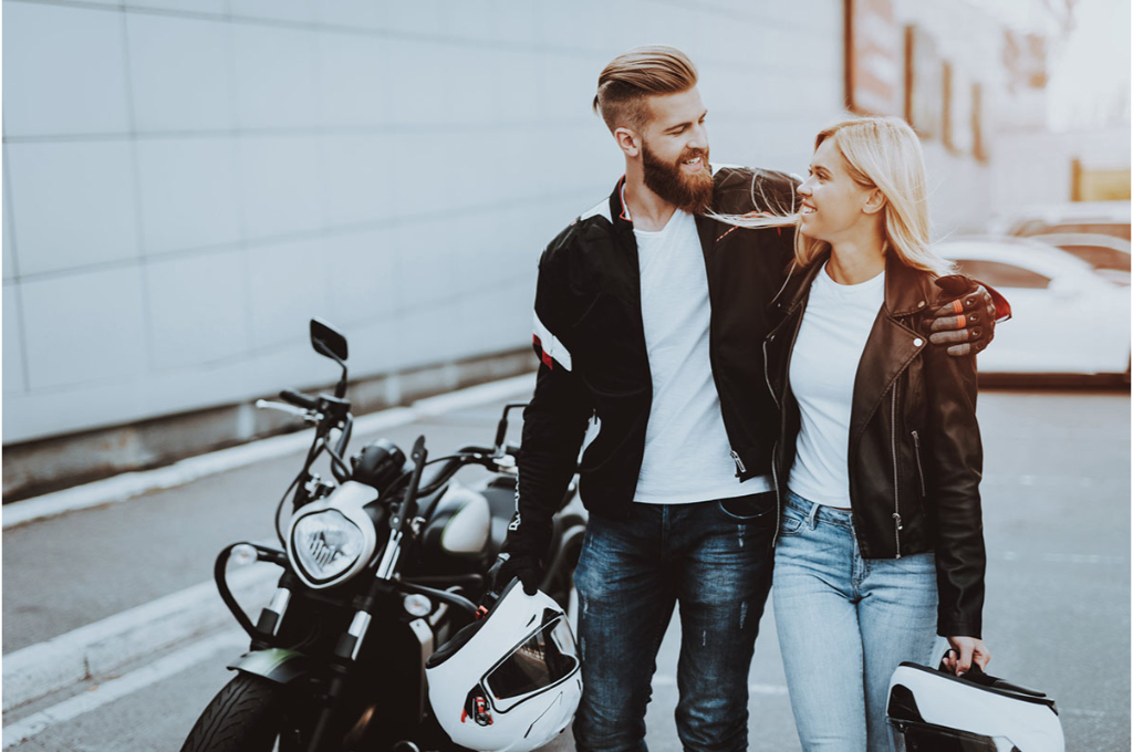 un couple se regarde en souriant devant une moto dans la rue. ils portent tous les deux une veste en cuir et leur casque de moto à la main.