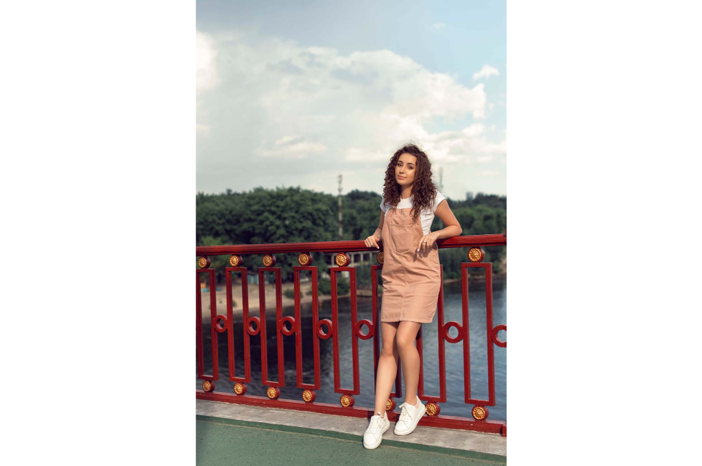 une jeune fille est adossée à la rambarde d'un pont dans une salopette robe courte, posée par-dessus un tee-shirt blanc. elle porte des baskets blanches.