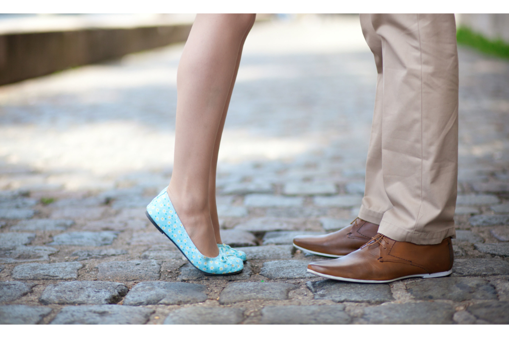 vue sur les jambes et chaussures de 2 personnes. La première personne est une femme en ballerines bleues à fleurs blanches. Elle est sur la pointe des pieds. La deuxième personne est un homme qui fait face à l'autre. Il porte des souliers en cuirs marrons et semelles blanches. Un pantalon beige tombe impeccablement sur ses chaussures.