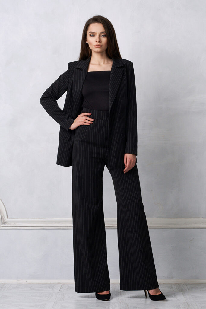 une femme pose en tailleur noir à rayures fines. elle porte des talons noirs.