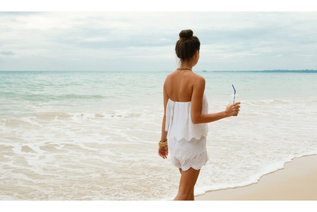 au bord de l'eau, sur une plage, une femme se promène en combi short blanc à dentelle. elle est de dos et tient une tasse en plastique avec une paille. elle a un chignon haut.