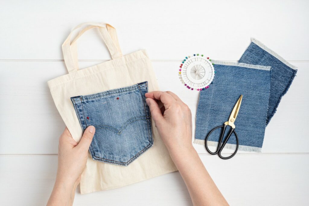 un tote bag est confectionné par des mains qui cousent une poche de jeans sur le tote bag. des chutes de jeans, aiguilles et ciseaux sont posés à côté.
