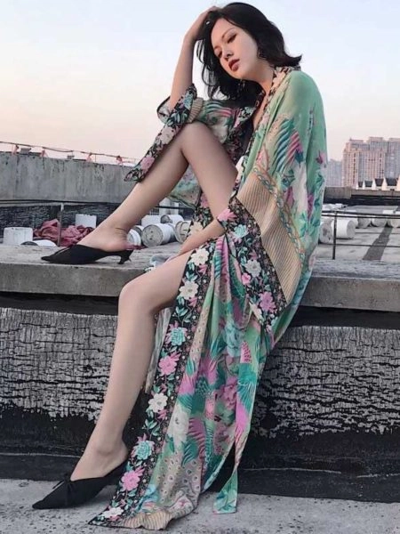 Une belle femme asiatique assise sur un mur avec une de ses jambes sur le mur porte un beau kimono style boho bohème longue fleuri vert à manches chauve-souris