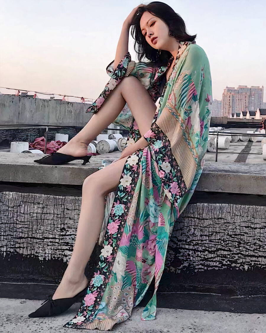 Une belle femme asiatique assise sur un mur avec une de ses jambes sur le mur porte un beau kimono style boho bohème longue fleuri vert à manches chauve-souris