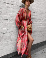 Une femme asiatique s'appuie sur un mur. Elle porte un chapeau large, do gros boucles d'oreille, un sac à main en cuir marron et un robe rouge fleuri en satin style kimono