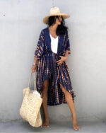 Femme contre un mur en beton, portant un short en jean avec un haut blanc et par dessus un kimono bleu foncé et un chapeau de paille, elle a un sac en osier de plage à la main droite et sa main gauche sur sa taille et elle regarde à gauche