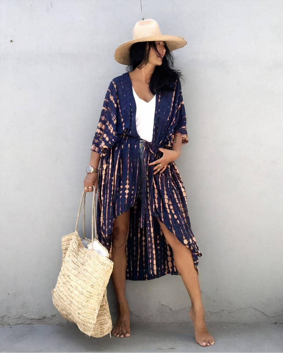 Femme contre un mur en beton, portant un short en jean avec un haut blanc et par dessus un kimono bleu foncé et un chapeau de paille, elle a un sac en osier de plage à la main droite et sa main gauche sur sa taille et elle regarde à gauche