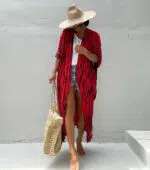 Femme qui porte un kimono de plage bohème rouge à pois noirs disposés en lignes verticales, qui regarde le sol et se tient de façon timide, elle tient un panier en osier à sa main droite, et porte un chapeau d'été