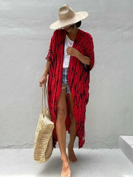 Femme qui porte un kimono de plage bohème rouge à pois noirs disposés en lignes verticales, qui regarde le sol et se tient de façon timide, elle tient un panier en osier à sa main droite, et porte un chapeau d'été