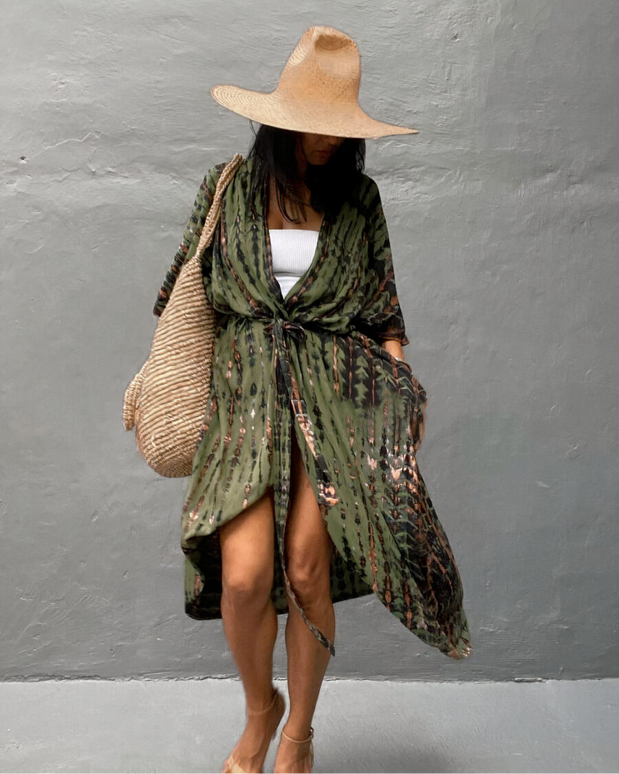 Femme qui porte u haut bas et par dessus un kimono bohème vert , noué à la taille , à l'épaule droite elle porte un grand sac de plage en osier beige et elle a aussi un chapeau en paille , sa tête est baissée vers le bas