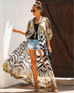 Femme qui se tient debout en talon hauts, devant un volet en bois marron, qui porte un short en jean court, un haut noir, et un kimono de plage aux motifs zébré noir et blanc, elle porte aussi des lunettes et ses cheveux sont attachés en chignon haut