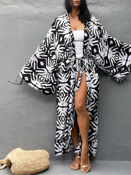 Dos à un mur gris, une femme brune, porte un kimono de plage long avec larges manches longues à imprimé bohème noir et blanc, en dessou du quel elle a un maillot blanc, et des talons hauts, ses bras sont ouvert vers le bas, presque en crois, et son sac de plage beige est jeté au sol