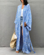 Devant un mur gris en beton, une femme brune se tient debout , elle porte un sac à man beige de plage dans sa main droite, elle porte également un short en jean court, un tee-shirt blanc et par dessus un kimono de plage aux imprimés léopard bleu à manche longue et bouffante