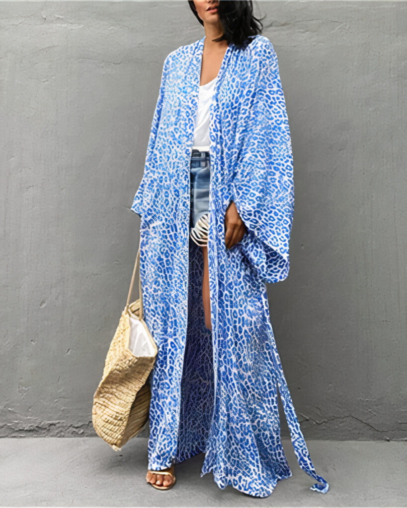 Devant un mur gris en beton, une femme brune se tient debout , elle porte un sac à man beige de plage dans sa main droite, elle porte également un short en jean court, un tee-shirt blanc et par dessus un kimono de plage aux imprimés léopard bleu à manche longue et bouffante