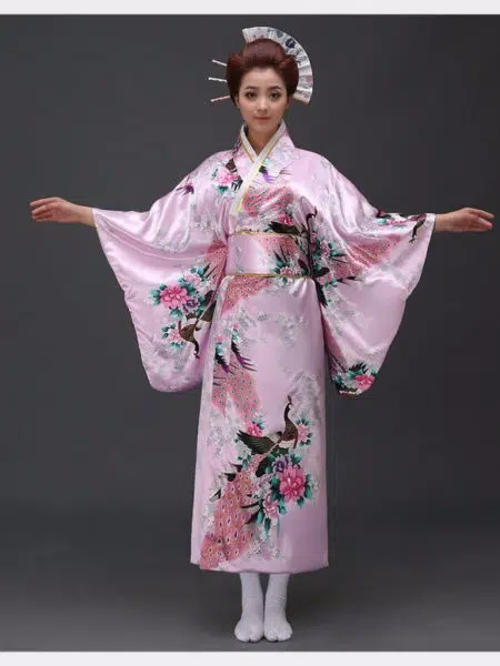 Une femme porte un kimono rose pastel avec des motifs japonais à manches larges, ses cheveux sont coiffés en chignons traditionnels, ses bras son ouverts de part et d'autre