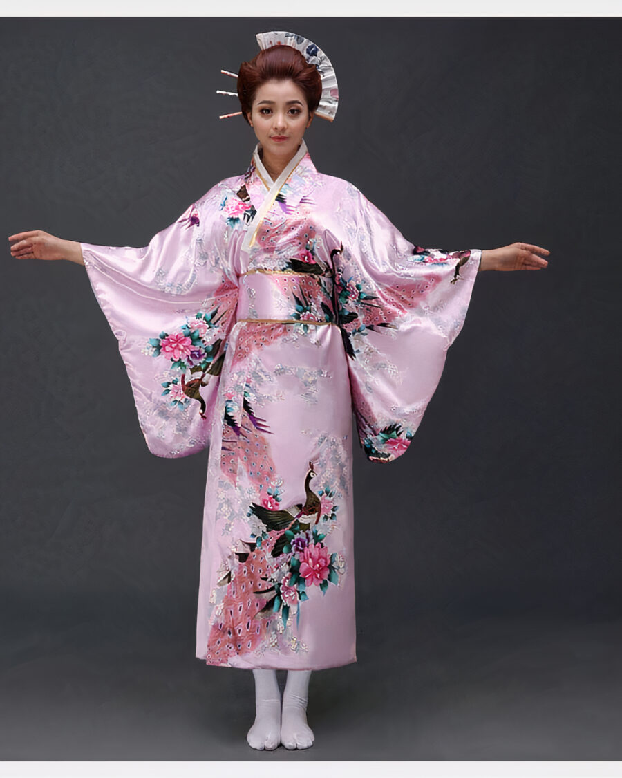 kimono-japonais-pour-femme Une femme porte un kimono rose pastel avec des motifs japonais à manches larges, ses cheveux sont coiffés en chignons traditionnels, ses bras son ouverts de part et d'autre
