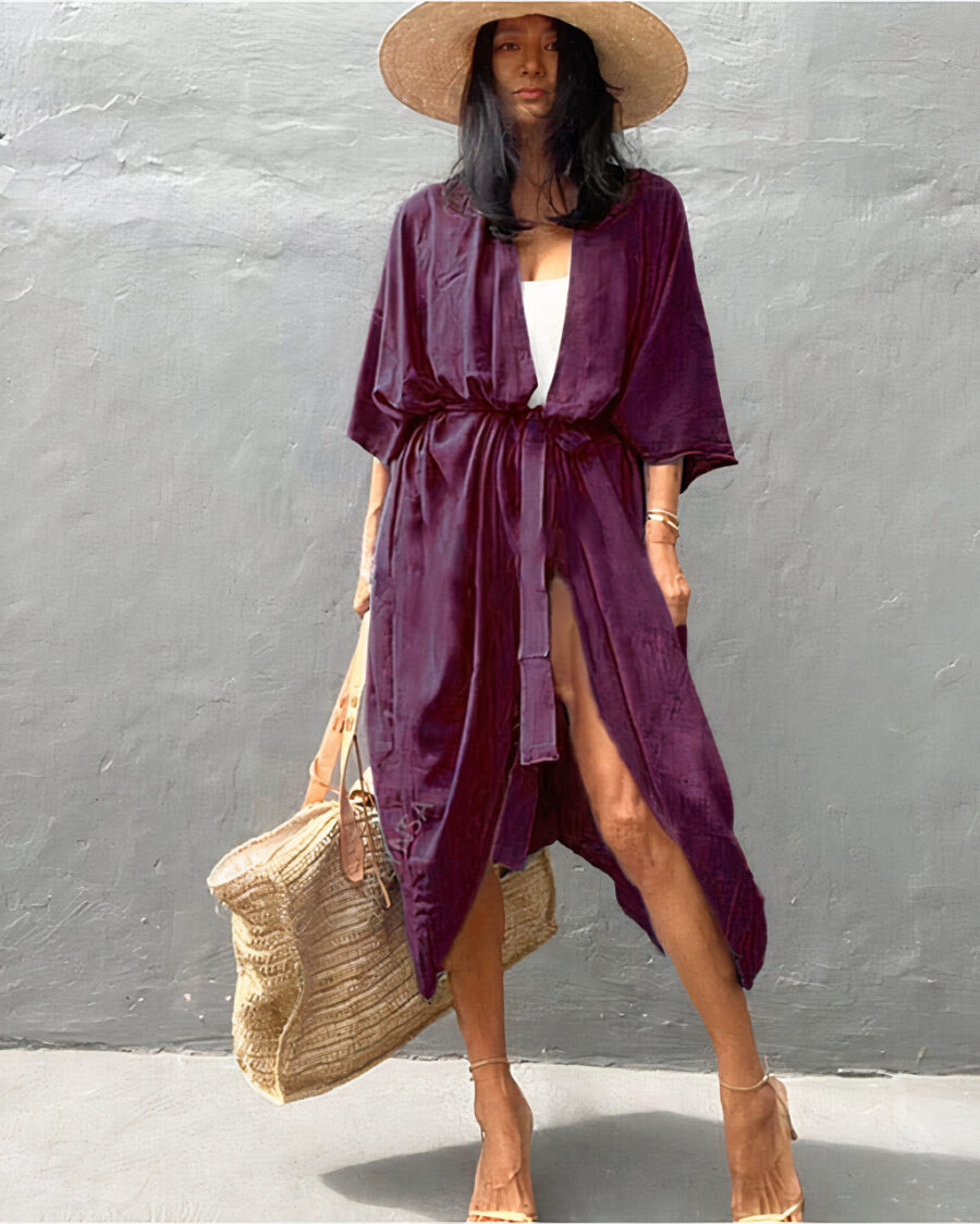 Femme brune debout contre un mur, porte un maillot blanc et par dessus lequel elle porte un kimono long violet,elle porte un sac de plage beige dans sa main droite, et un chapeau en paille