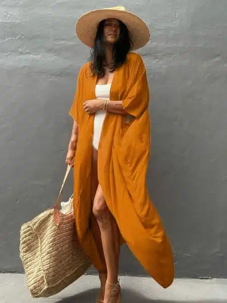 Femme brune debout contre un mur, porte un maillot blanc et par dessus lequel elle porte un kimono long orange,elle porte un sac de plage beige dans sa main droite, et un chapeau en paille