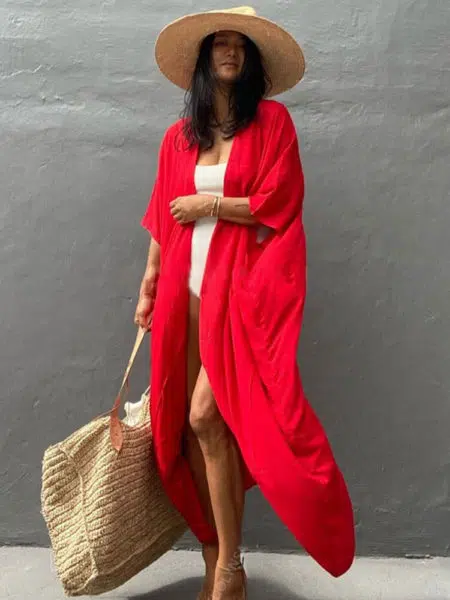 Femme brune debout contre un mur, porte un maillot blanc et par dessus lequel elle porte un kimono long rouge, elle tient son sac de plage beige dans sa main droite le long de son corps