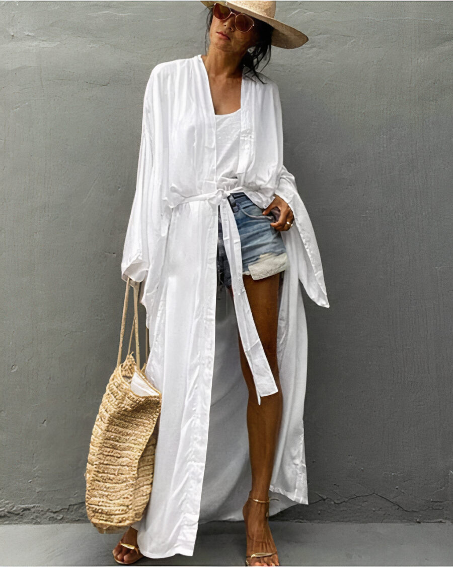 Kimono blanc porté par une femme avec un chapeau et un sac à main en paille, un haut blanc et un short en jean et des sandales dorées devant un mur gris