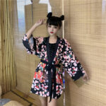 Une femme debout dans une chambre portant un kimono de style japonais de couleur noir et rose avec des motifs fleuris colorés. Le kimono est fermé par une ceinture noire.