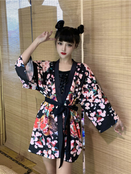 Une femme debout dans une chambre portant un kimono de style japonais de couleur noir et rose avec des motifs fleuris colorés. Le kimono est fermé par une ceinture noire.