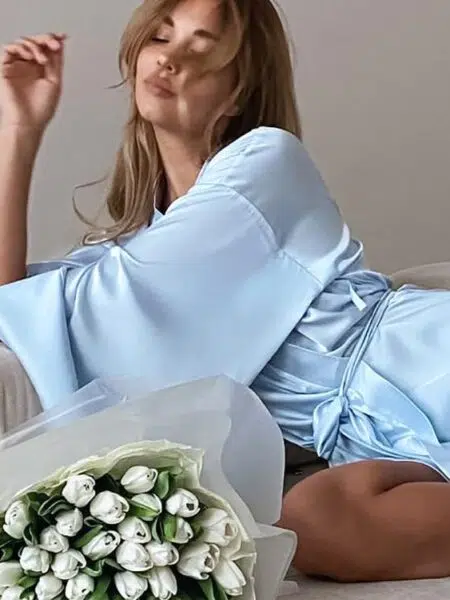 installée à demi allongée, une femme se trouve sur un canapé et porte un peignoir kimono bleu clair et près d'elle des fleurs blanches en bouquet
